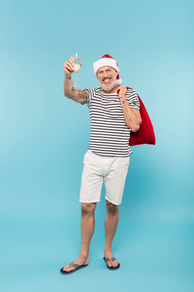 plná délka šťastného muže středního věku v Santa klobouku drží pytel a sklenici šampaňského na modré - Fotografie, Obrázek