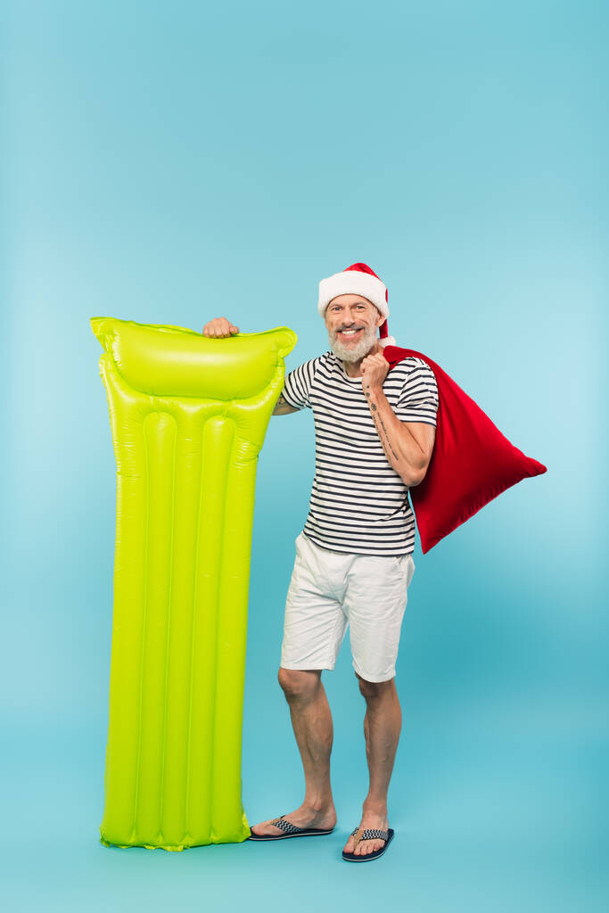 plná délka šťastný muž středního věku v Santa klobouk drží pytel a nafukovací matrace na modré - Fotografie, Obrázek