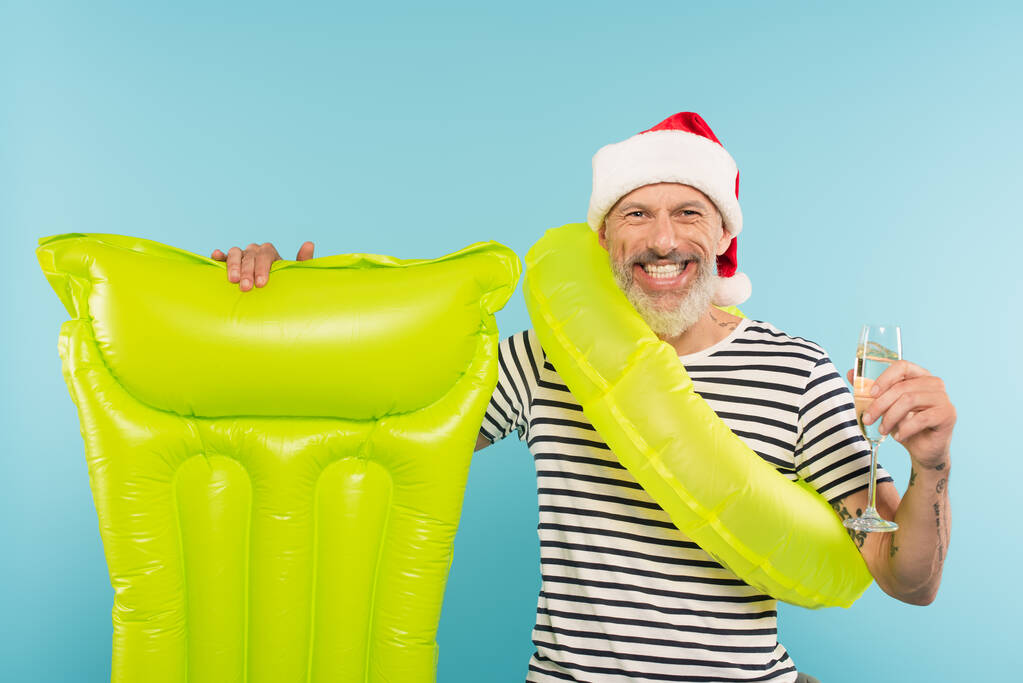 happy middle aged man in santa hat with swim ring standing near inflatable mattress and holding glass of champagne isolated on blue - Photo, Image