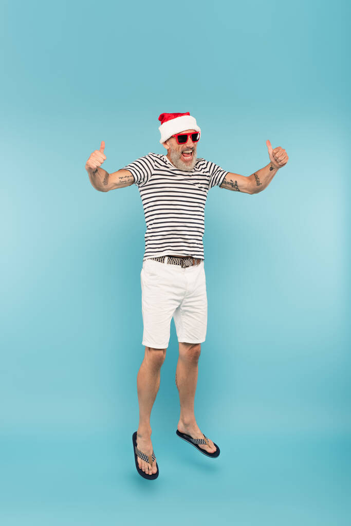 in voller Länge glücklicher Mann mit Sonnenbrille und Weihnachtsmütze springt und zeigt Daumen hoch auf blau - Foto, Bild