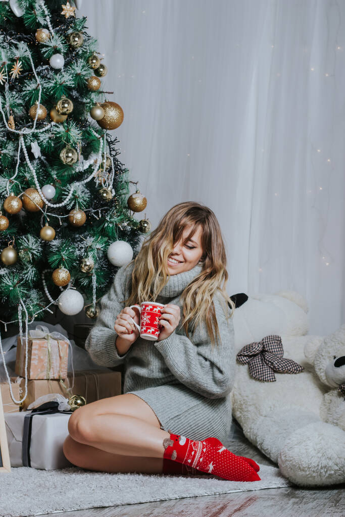 Glückliche blonde Frau sitzt auf dem Boden, hält einen roten Becher mit Hirsch in der Hand und trinkt Kaffee oder Tee auf dem Hintergrund des geschmückten Weihnachtsbaums. Die Atmosphäre des Urlaubs - Foto, Bild