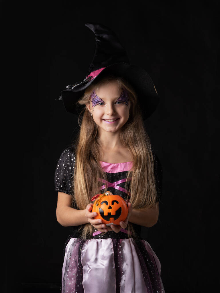 Милая девочка-ведьма на Хэллоуин в сказочном костюме, держит ведро с тыквой и леденцами, изолированные на черном фоне - Фото, изображение