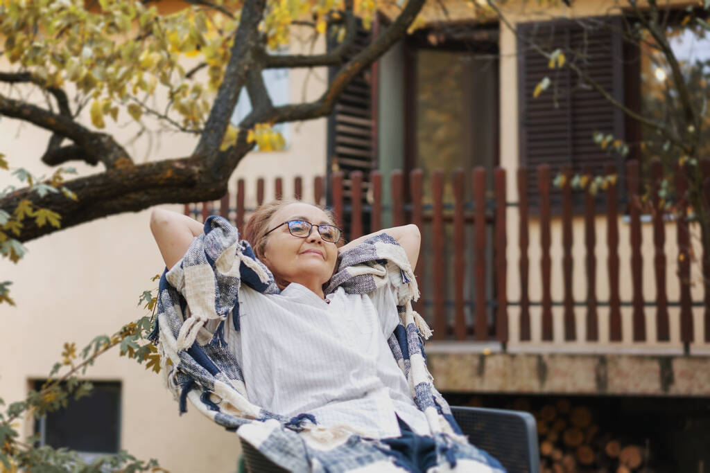 Mooie volwassen gelukkig glimlachende vrouw ontspannen terwijl zitten in een fauteuil in de herfst tuin van haar landhuis - Foto, afbeelding