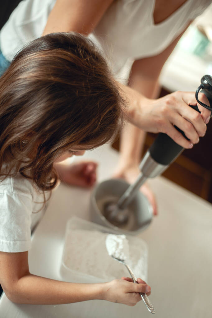 Η μαμά και η κόρη ετοιμάζουν γλάσο για μελόψωμο στην κουζίνα του σπιτιού τους. Χτυπάμε με ένα μπλέντερ. Το κορίτσι βοηθάει τη γυναίκα.. - Φωτογραφία, εικόνα