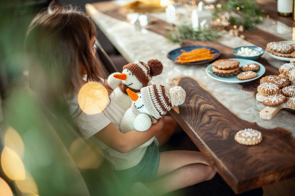 Egy aranyos kislány ül egy asztalnál, tele karácsonyi dekorációval. És két kötött hóembert tart a kezében.. - Fotó, kép