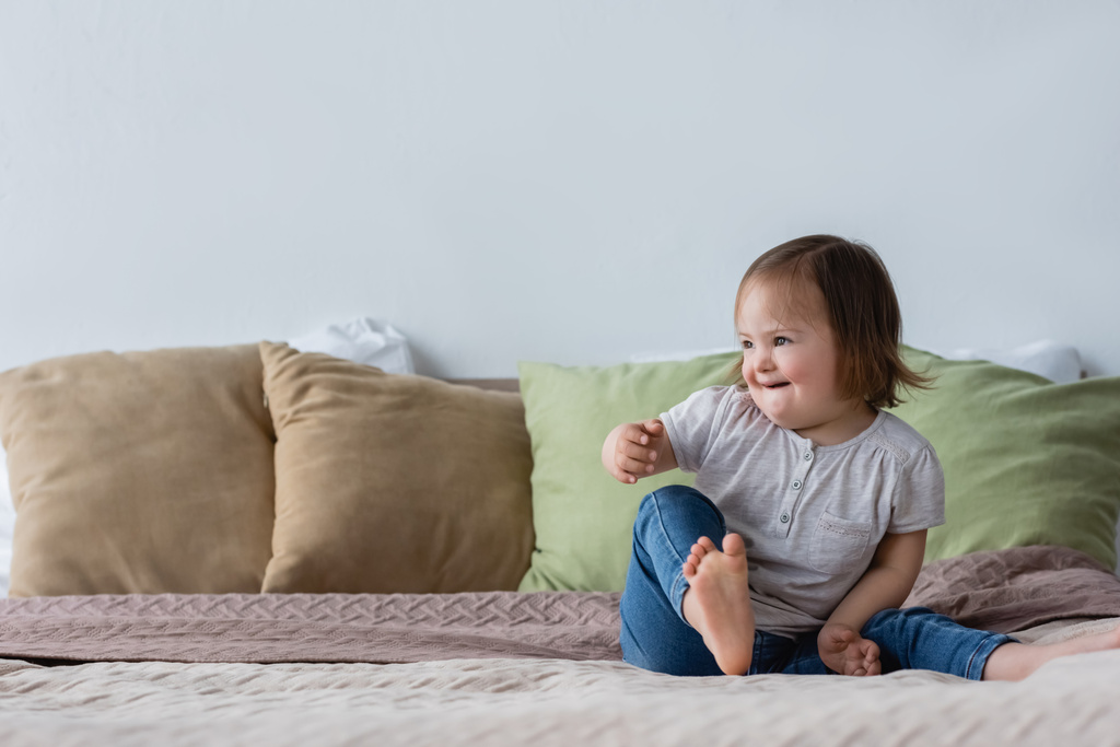 Mosolygós és mezítlábas gyerek Down-szindrómával az ágyon ülve  - Fotó, kép