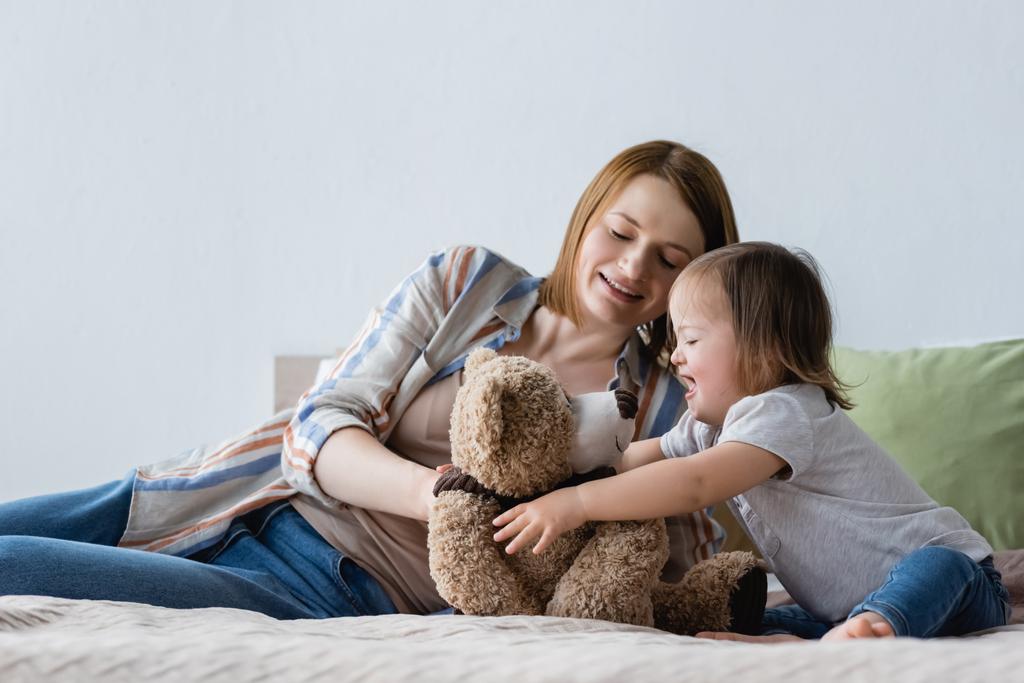 Усміхнена жінка грає з дочкою з синдромом вниз і м'яка іграшка на ліжку
  - Фото, зображення