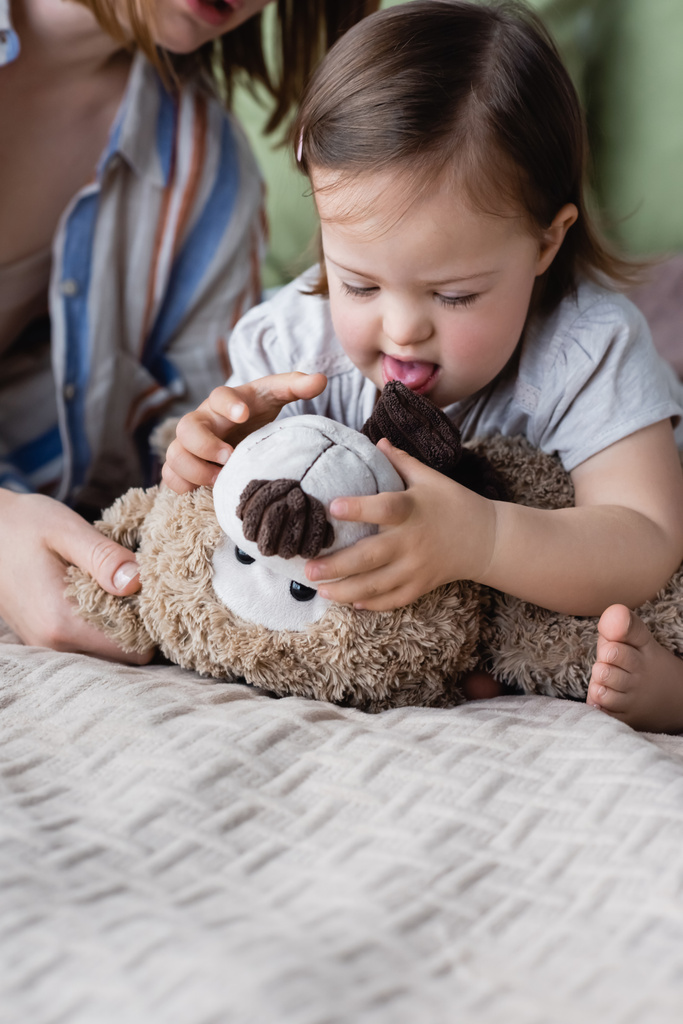 Niña con síndrome de Down sosteniendo juguete suave cerca de la madre borrosa en la cama  - Foto, Imagen