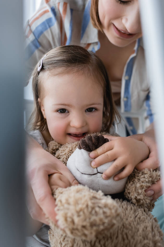 Pozitivní dítě s Downovým syndromem drží měkkou hračku v blízkosti usmívající se matky v postýlce  - Fotografie, Obrázek
