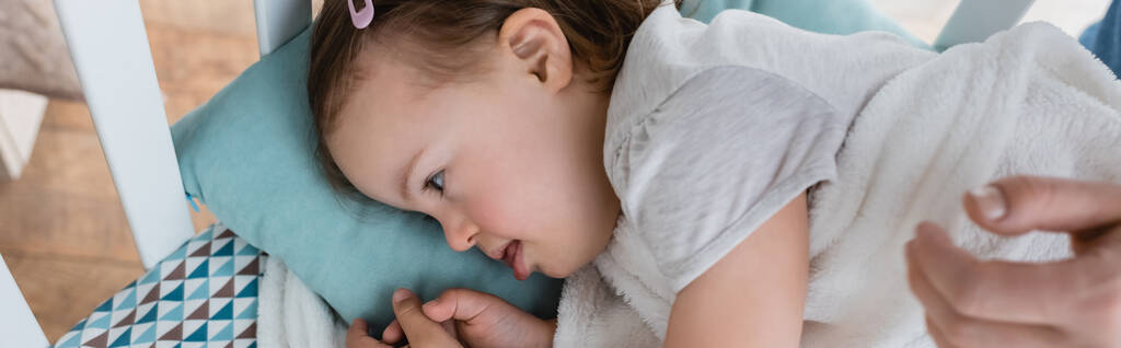 Widok boczny dziecka z zespołem Downa leżącego w łóżeczku obok ręki matki, sztandar  - Zdjęcie, obraz