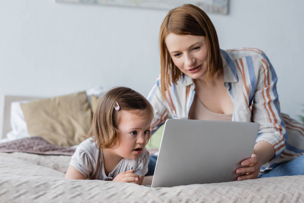 Bambino con sindrome di Down guardando computer portatile vicino alla madre offuscata in camera da letto  - Foto, immagini