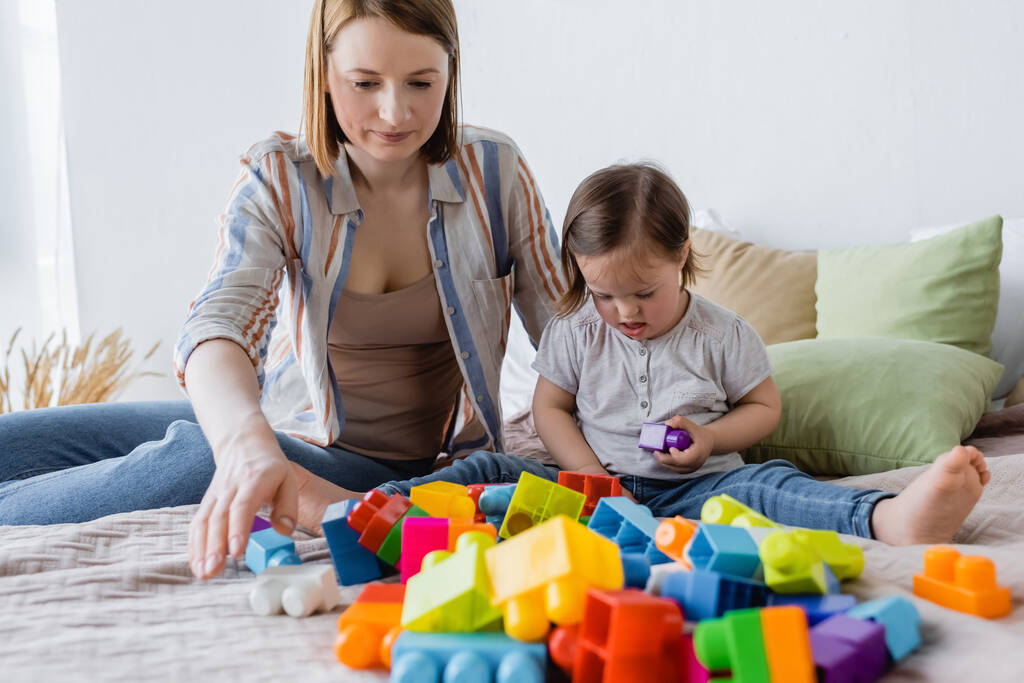 Down sendromlu ebeveyn ve çocuk evde yatakta renkli yapı taşları oynuyorlar.  - Fotoğraf, Görsel