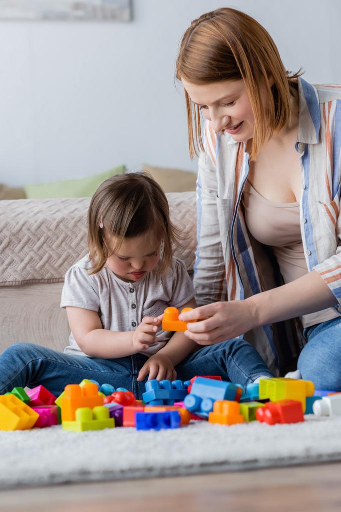 Femme jouant bloc de construction avec l'enfant avec le syndrome du duvet sur le tapis dans la chambre  - Photo, image
