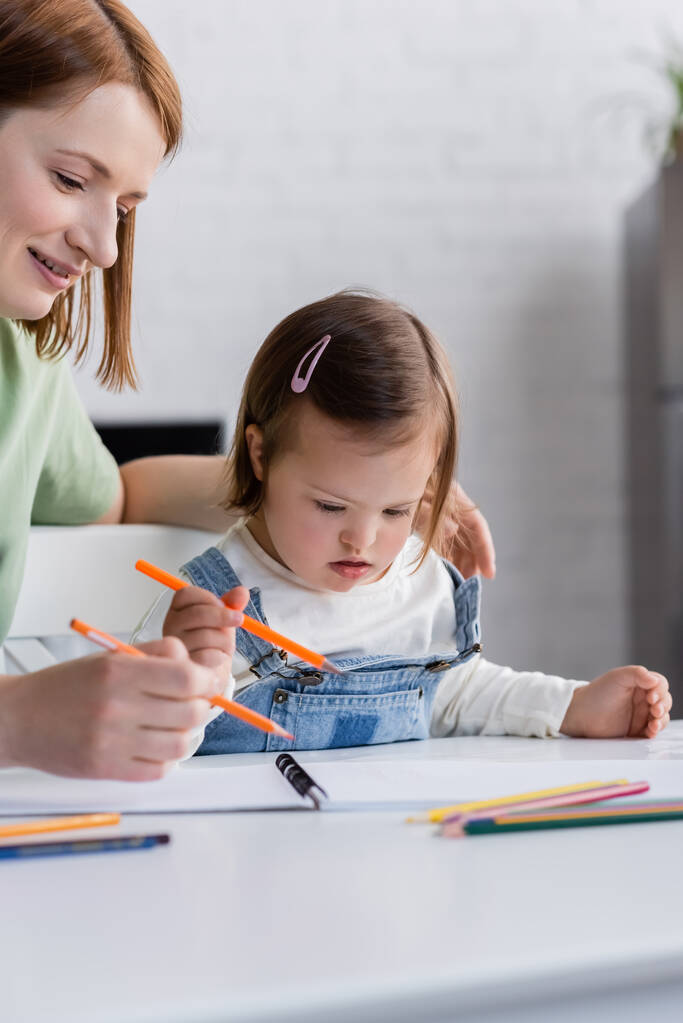 Femme souriante tenant crayon de couleur près de la fille avec le syndrome du duvet et du papier à la maison  - Photo, image