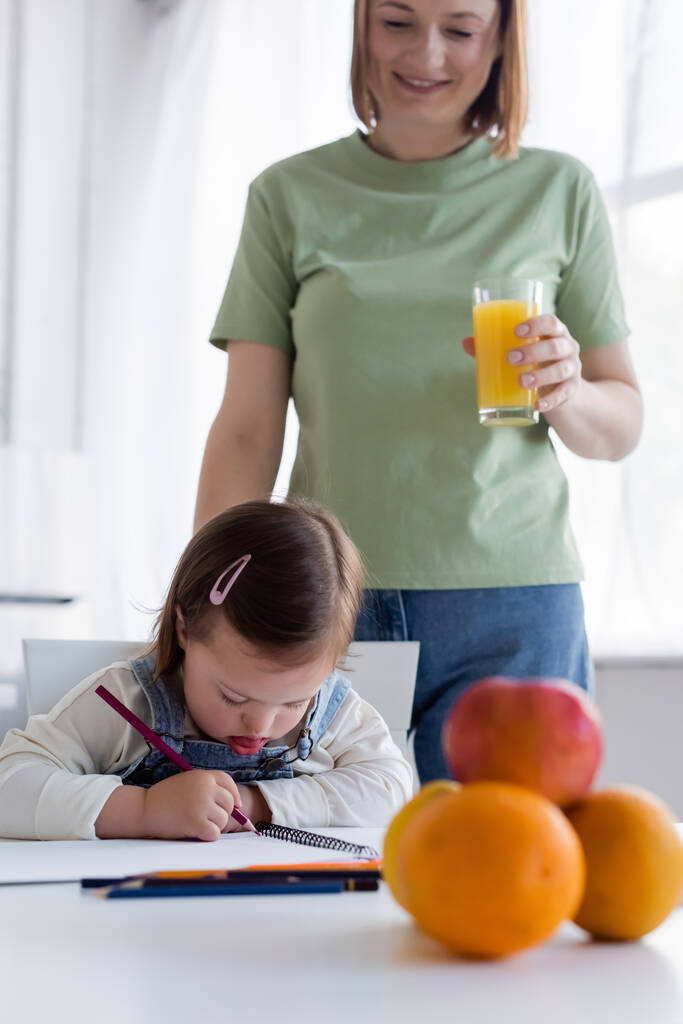 Dzieciak z zespołem Downa rysunek blisko uśmiechnięta mama z sokiem pomarańczowym w kuchni  - Zdjęcie, obraz