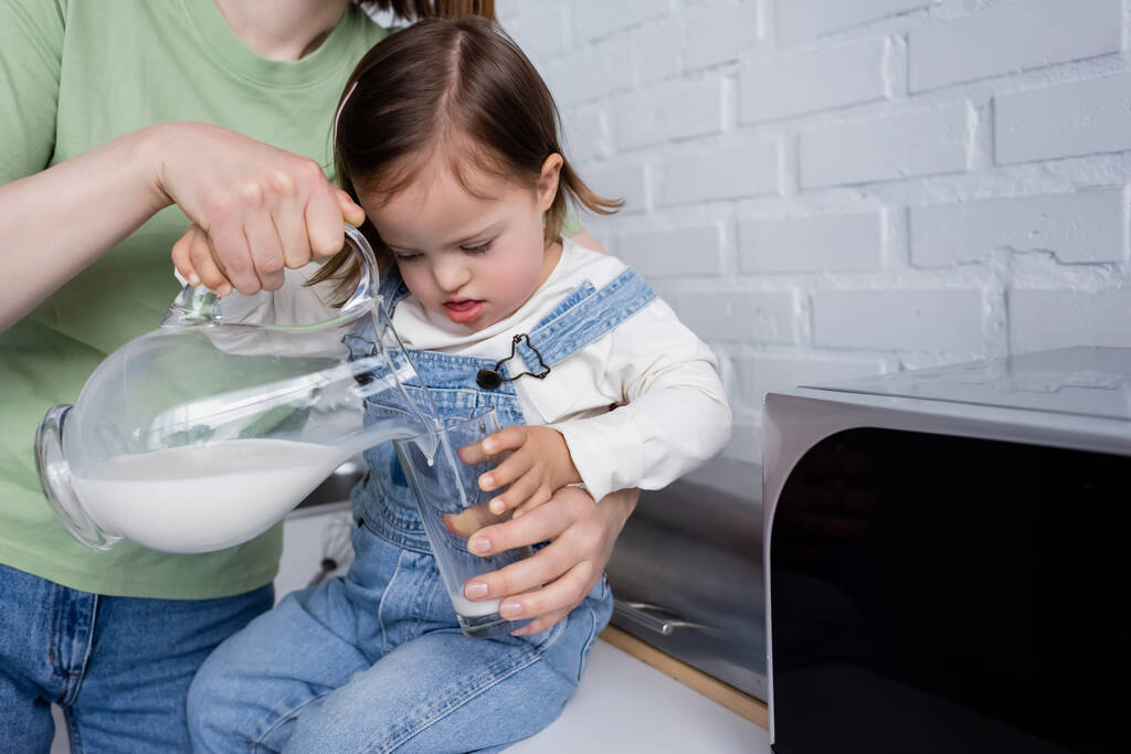 Kobieta wylewa mleko do szklanki w pobliżu córki z zespołem Downa w kuchni  - Zdjęcie, obraz