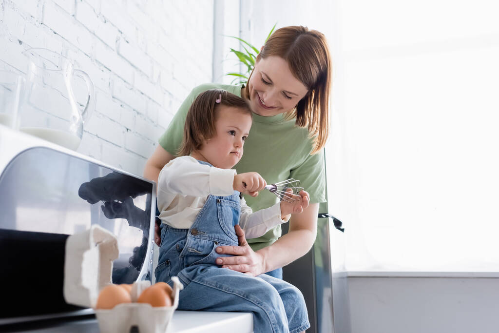 Счастливая мать, стоящая рядом с ребенком с синдромом Дауна и ингредиентами на кухне  - Фото, изображение