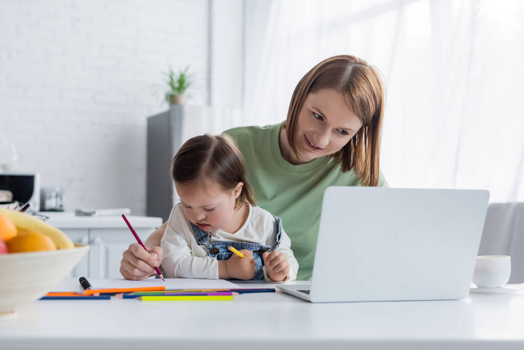 Freelance sorridente che guarda il computer portatile vicino a figlia con sindrome giù che disegna in cucina  - Foto, immagini