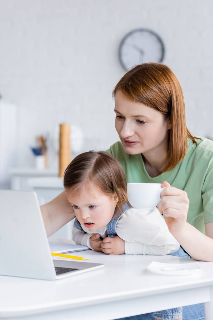Woman holding cup near daughter with down syndrome and laptop in kitchen  - Photo, Image
