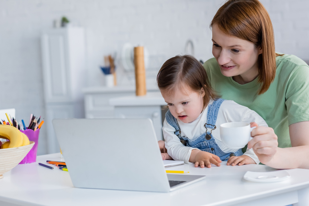 Gülümseyen kadın, elinde bardakla aşağı sendromlu bir çocuk, dizüstü bilgisayar ve renkli kalemler tutuyor mutfakta.  - Fotoğraf, Görsel