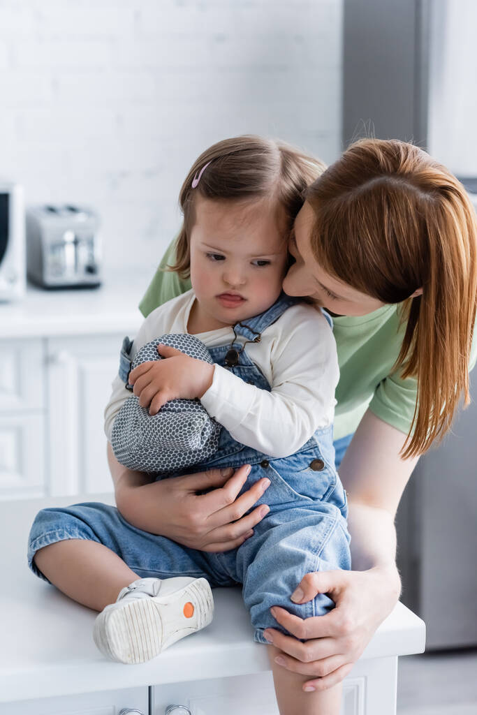 Mutter umarmt Tochter mit Down-Syndrom, streckt Zunge heraus und hält Backhandschuh in Küche  - Foto, Bild