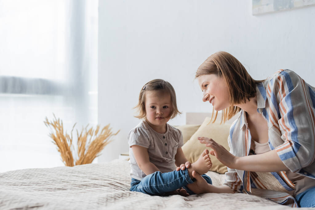 Улыбающаяся мать касается ног маленькой дочери с синдромом Дауна на кровати  - Фото, изображение