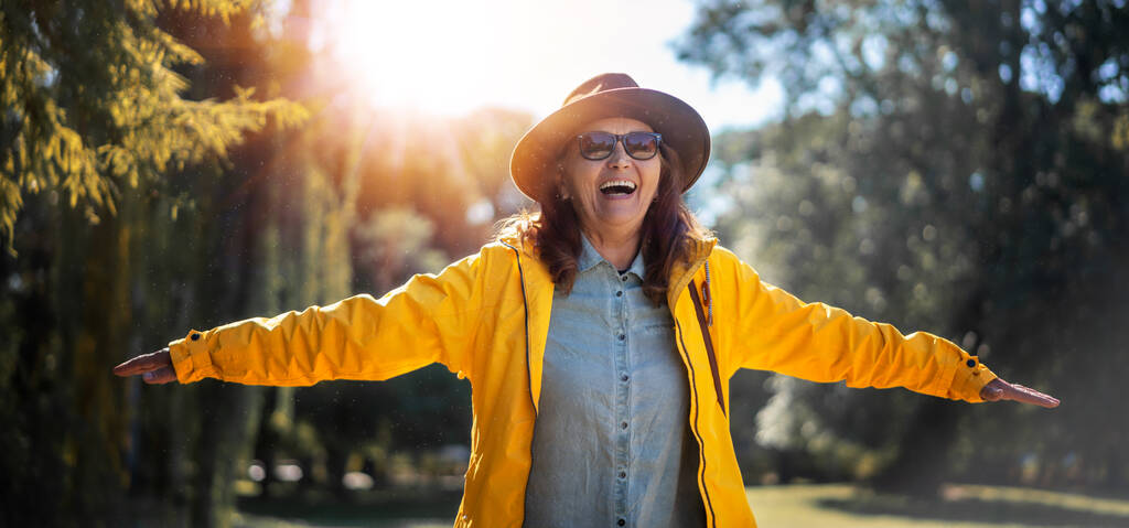 美しいです幸せな成熟した女性で帽子と黄色のジャケット楽しんで自然で公園ストレッチ彼女の腕 - 写真・画像