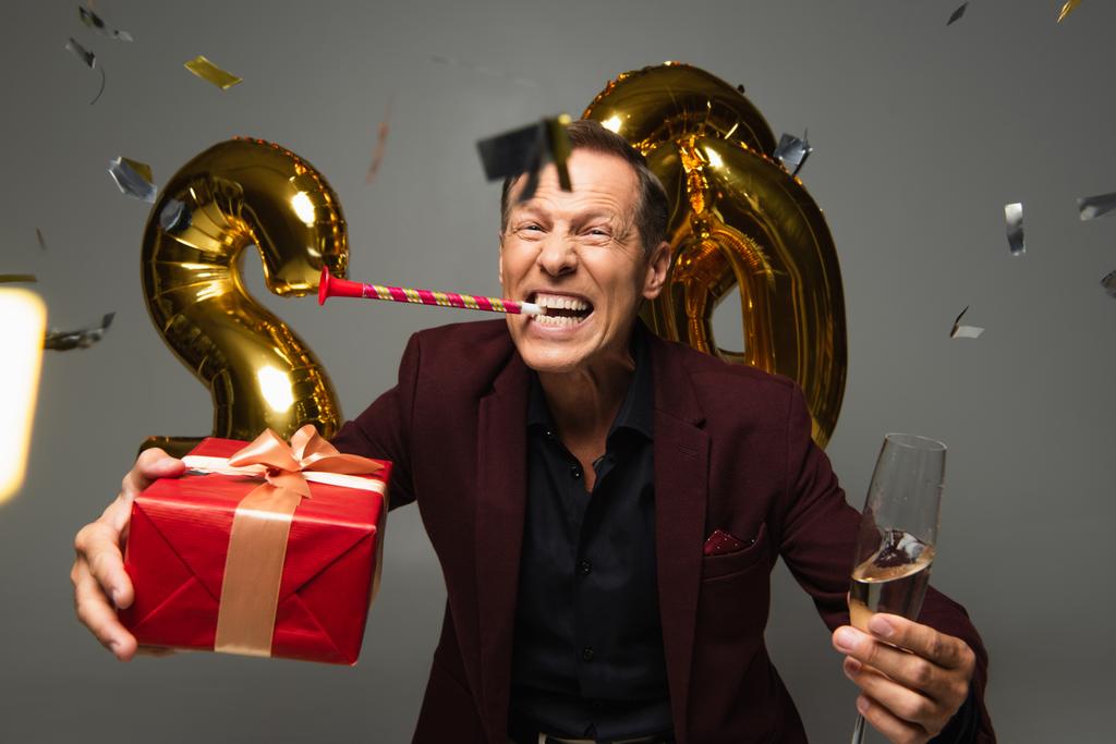 Vzrušený dospělý muž s party rohu drží dárkové krabice a šampaňské v blízkosti balónky a konfety na šedém pozadí  - Fotografie, Obrázek