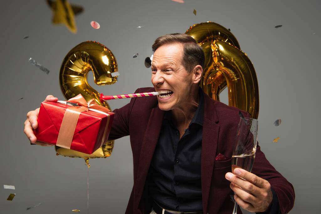 Parti boynuzu taşıyan heyecanlı adam balonların yanında şampanya ve gri arka planda konfeti tutuyor.  - Fotoğraf, Görsel