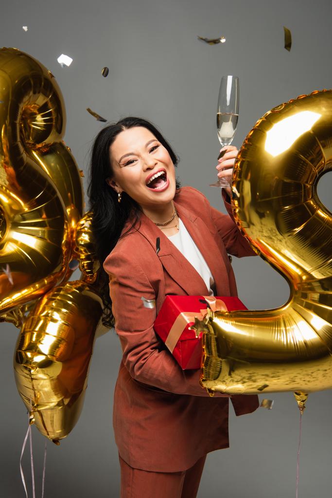 Emocionado mujer asiática sosteniendo copa de champán y presente cerca de confeti y globos sobre fondo gris  - Foto, imagen