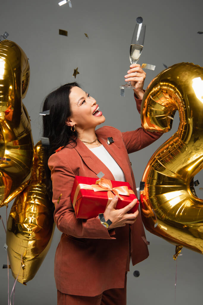 Femme asiatique excitée avec cadeau regardant un verre de champagne près de ballons et confettis sur fond gris  - Photo, image