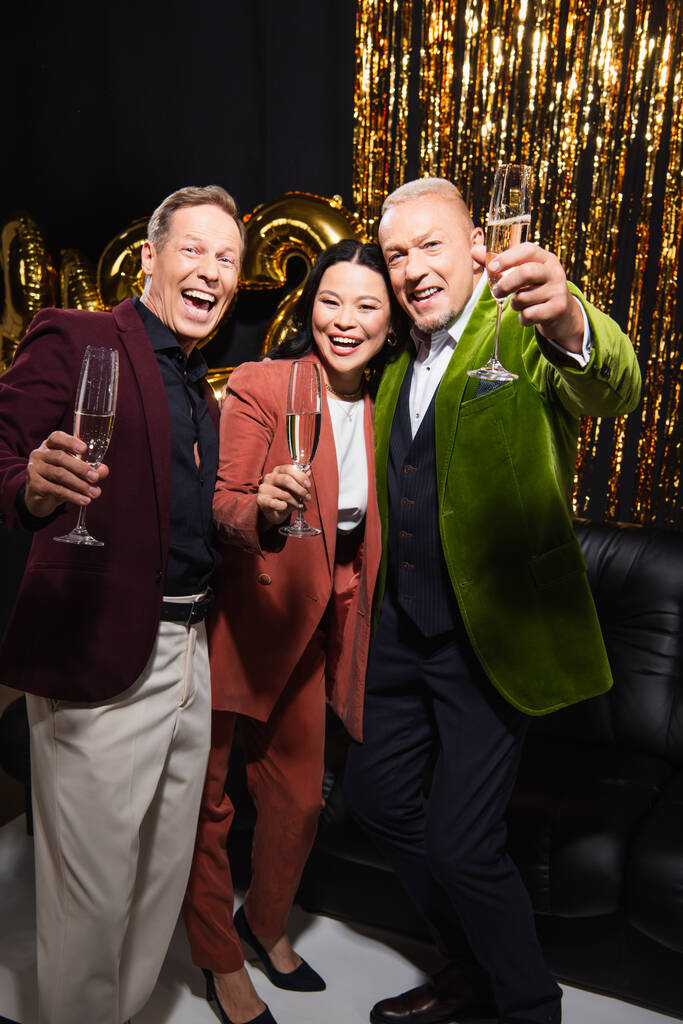 Happy interracial starší přátelé drží šampaňské a při pohledu na kameru v blízkosti balónky a tinsel na černém pozadí  - Fotografie, Obrázek