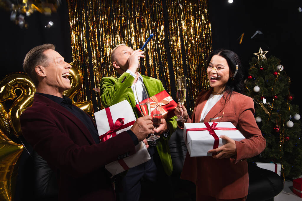 Felices amigos multiétnicos con champán, regalos y cuerno de fiesta celebrando el año nuevo sobre fondo negro  - Foto, Imagen