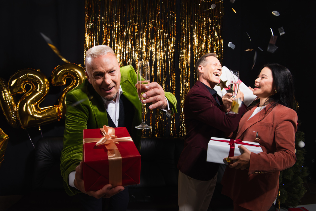 Positieve volwassen man met champagne en presenteren in de buurt van interraciale vrienden tijdens nieuwjaar feest op zwarte achtergrond  - Foto, afbeelding
