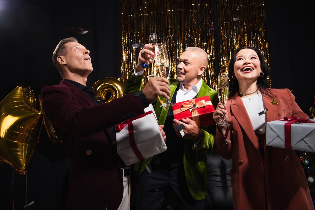 Lächelnd interrassische reife Freunde mit Champagner und Geschenken während der Weihnachtsfeier auf schwarzem Hintergrund  - Foto, Bild