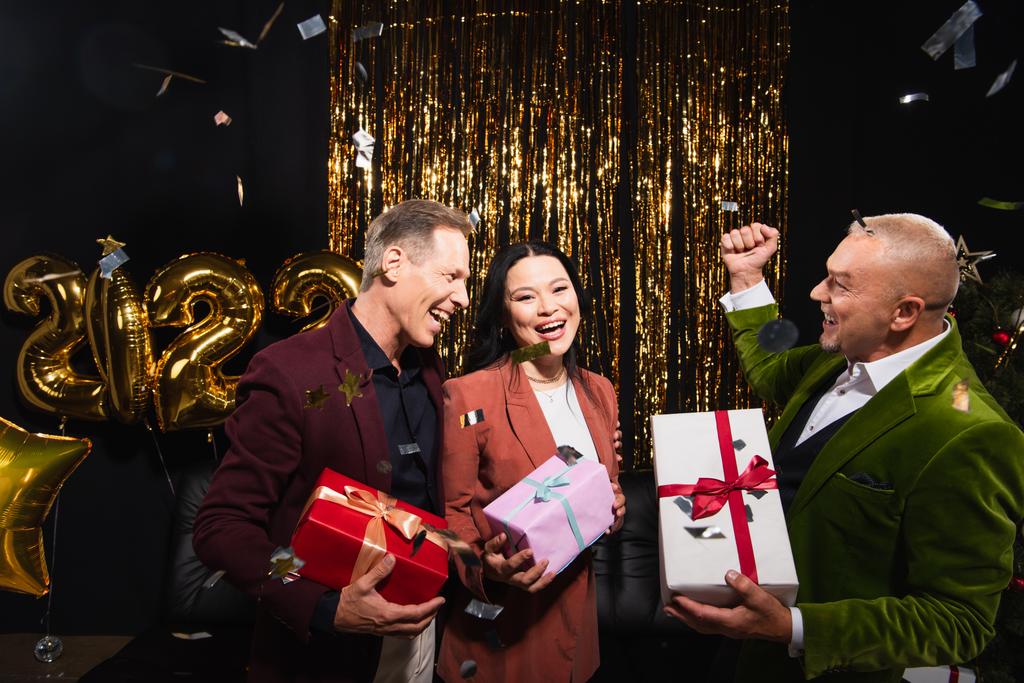 Emocionados amigos interracial con regalos celebrando año nuevo cerca de confeti y globos sobre fondo negro  - Foto, Imagen