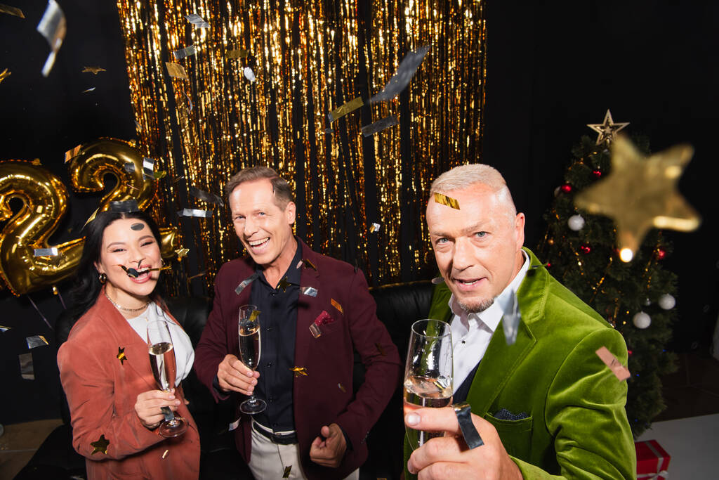 Высокоугольный вид зрелых межрасовых друзей с шампанским, смотрящих на камеру под конфетти во время новогодней вечеринки на черном фоне  - Фото, изображение
