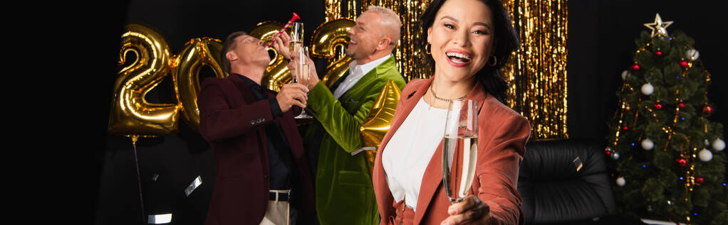 glimlachende aziatische vrouw met glas champagne kijken naar camera in de buurt van vrienden vieren van Nieuwjaar op zwarte achtergrond, banner   - Foto, afbeelding