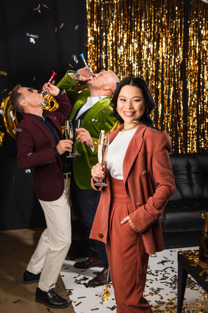 aziatische vrouw met champagne kijken naar camera in de buurt van vrienden met partij hoorn tijdens nieuwjaar partij op zwart achtergrond  - Foto, afbeelding