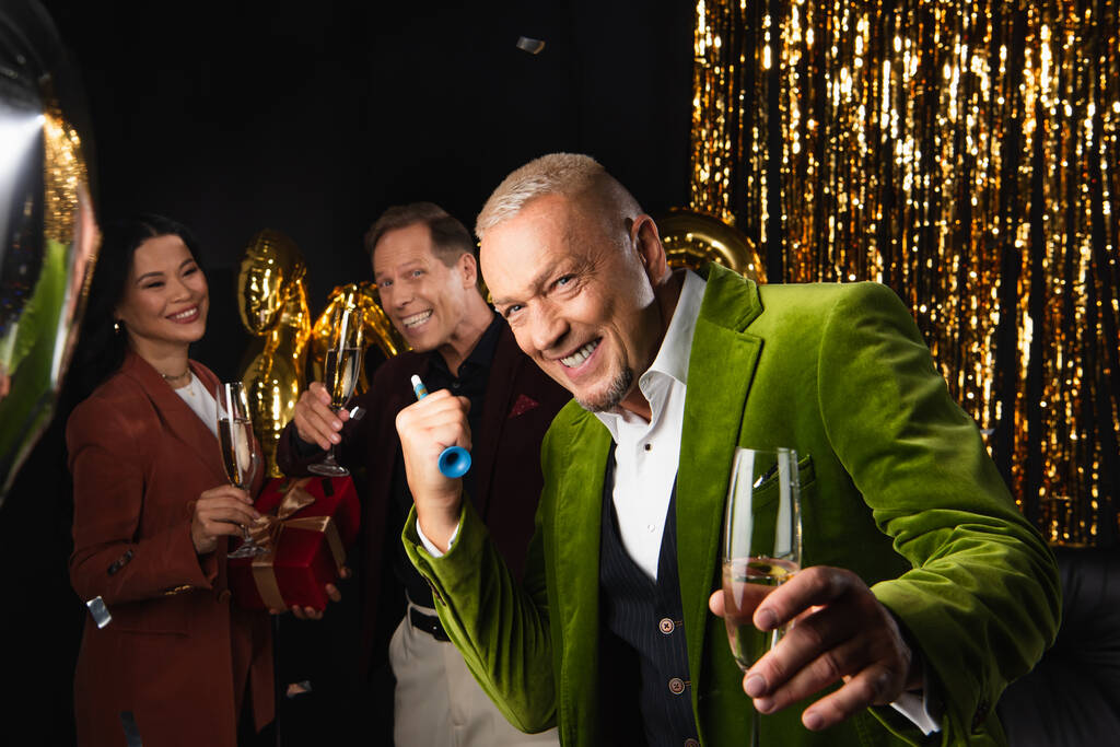 Homem maduro feliz segurando chifre de festa e champanhe perto de amigos multiétnicos e decoração festiva no fundo preto  - Foto, Imagem