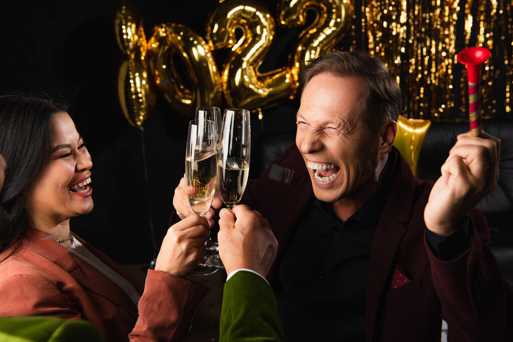 Позитивный человек с рогом для вечеринки, пьющий шампанское с многонациональными друзьями во время новогодней вечеринки на черном фоне  - Фото, изображение