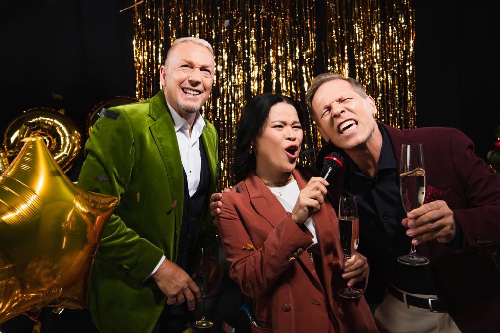 Mulher asiática com champanhe cantando karaoke com amigos maduros durante a festa no fundo preto  - Foto, Imagem