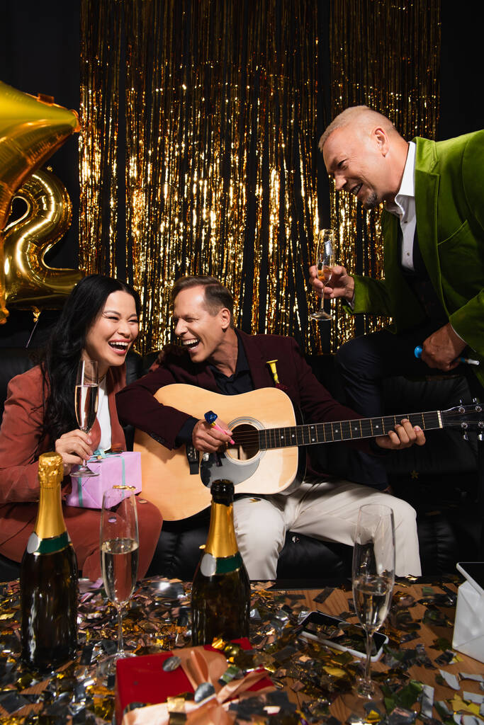Ami interracial positif avec champagne jouant de la guitare acoustique près de cadeaux lors de la fête du Nouvel An sur fond noir  - Photo, image