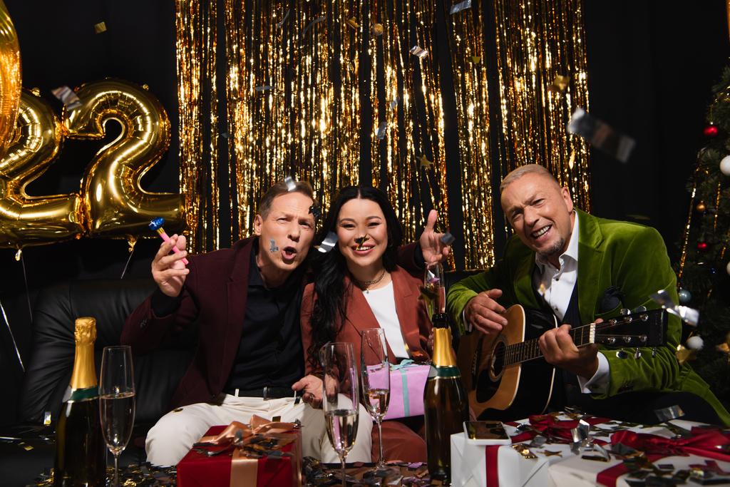Opgewonden interraciale vrienden met akoestische gitaar vieren nieuw jaar in de buurt van champagne en geschenken op zwarte achtergrond  - Foto, afbeelding