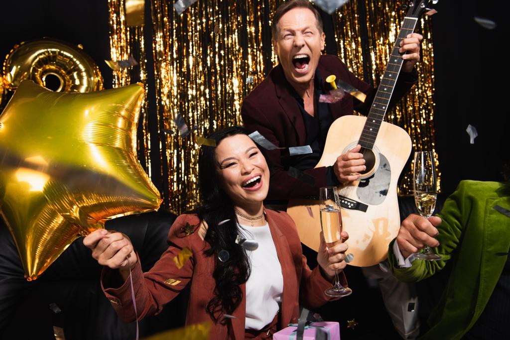 Balon ve şampanya tutan neşeli Asyalı kadın akustik gitarı ve siyah arka planda konfetisi olan arkadaşlarının yanında.  - Fotoğraf, Görsel