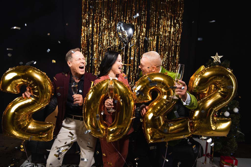 Podekscytowani wielonarodowymi przyjaciółmi z kieliszkami szampana świętującymi Nowy Rok w pobliżu balonów w kształcie 2022 roku na czarnym tle  - Zdjęcie, obraz