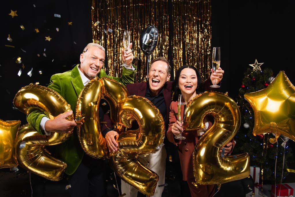 Cheerful multiethnic friends holding champagne near balloons in shape of 2022 during new year party on black background  - Photo, Image