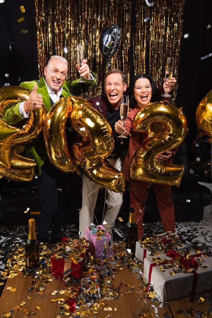 Ενθουσιασμένοι πολυεθνικοί φίλοι με σαμπάνια γιορτάζουν το νέο έτος κοντά σε κομφετί, δώρα και μπαλόνια σε μαύρο φόντο  - Φωτογραφία, εικόνα