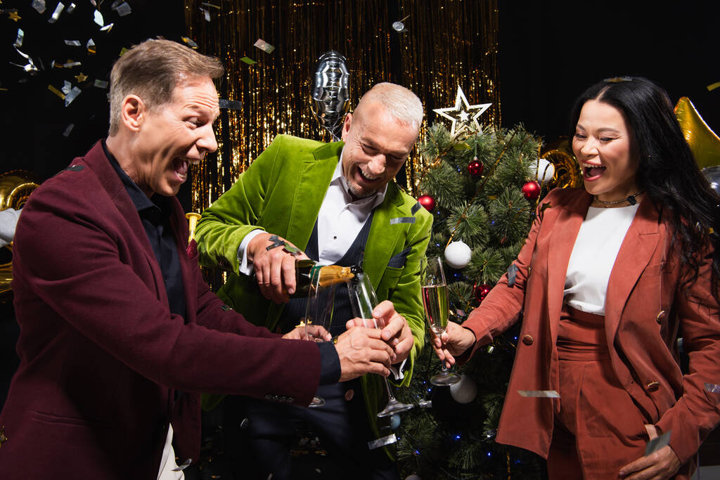 maturo uomo versando champagne vicino interrazziale amici e albero di Natale su sfondo nero  - Foto, immagini