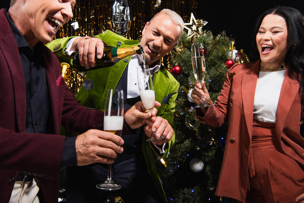 Mature man pouring champagne near excited interracial friends and christmas tree during new year party on black background  - Photo, Image